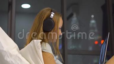 年轻女子戴着无线耳机，用笔记本电脑工作，她坐在床上，一个摩天大<strong>楼</strong>的<strong>剪影</strong>
